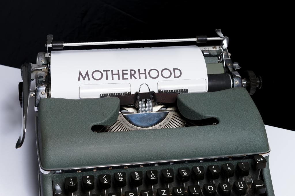 An old-fashioned typewriter with a piece of paper in it, displaying the word MOTHERHOOD in capital letters